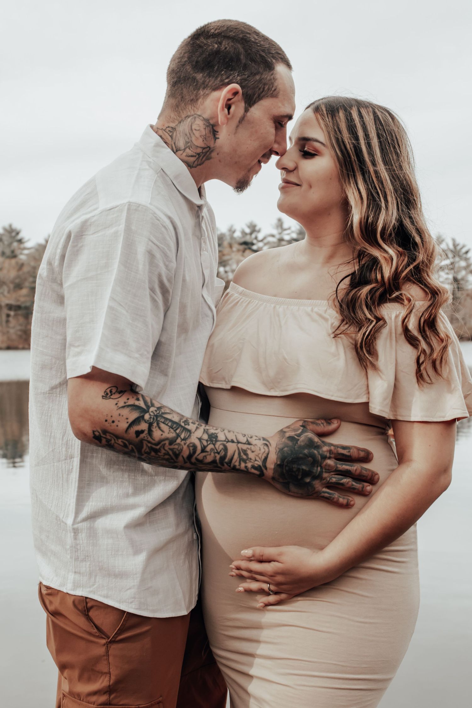 Intimate maternity portrait of couple in coordinated neutral outfits against lakeside backdrop.