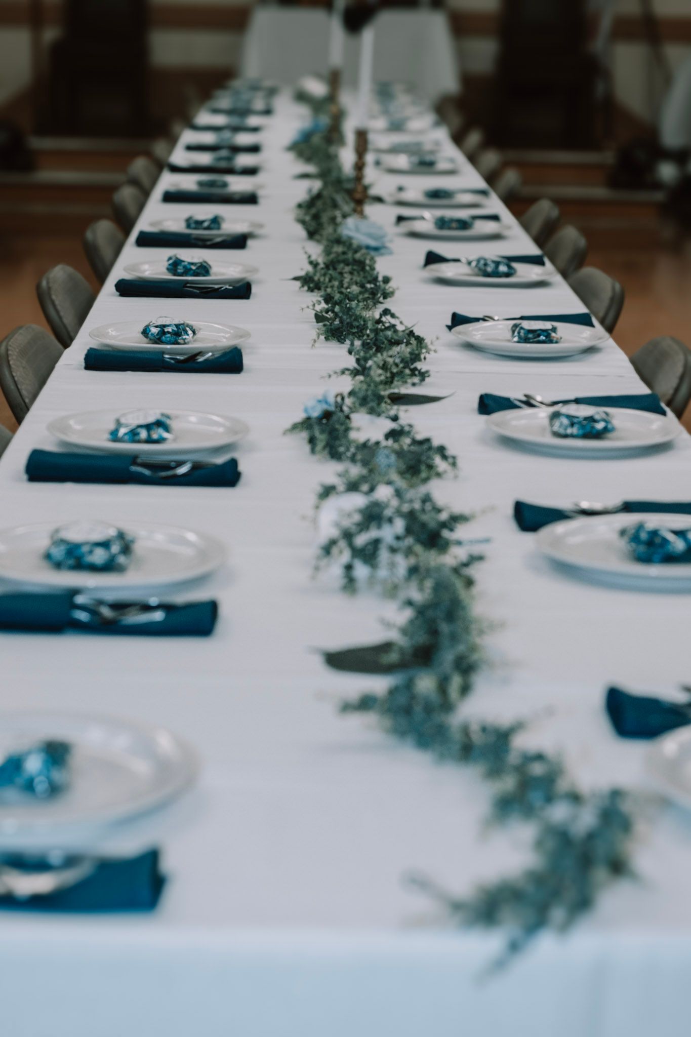 Elegant wedding table setting with white tablecloth, navy blue napkins and delicate greenery garland running down center.