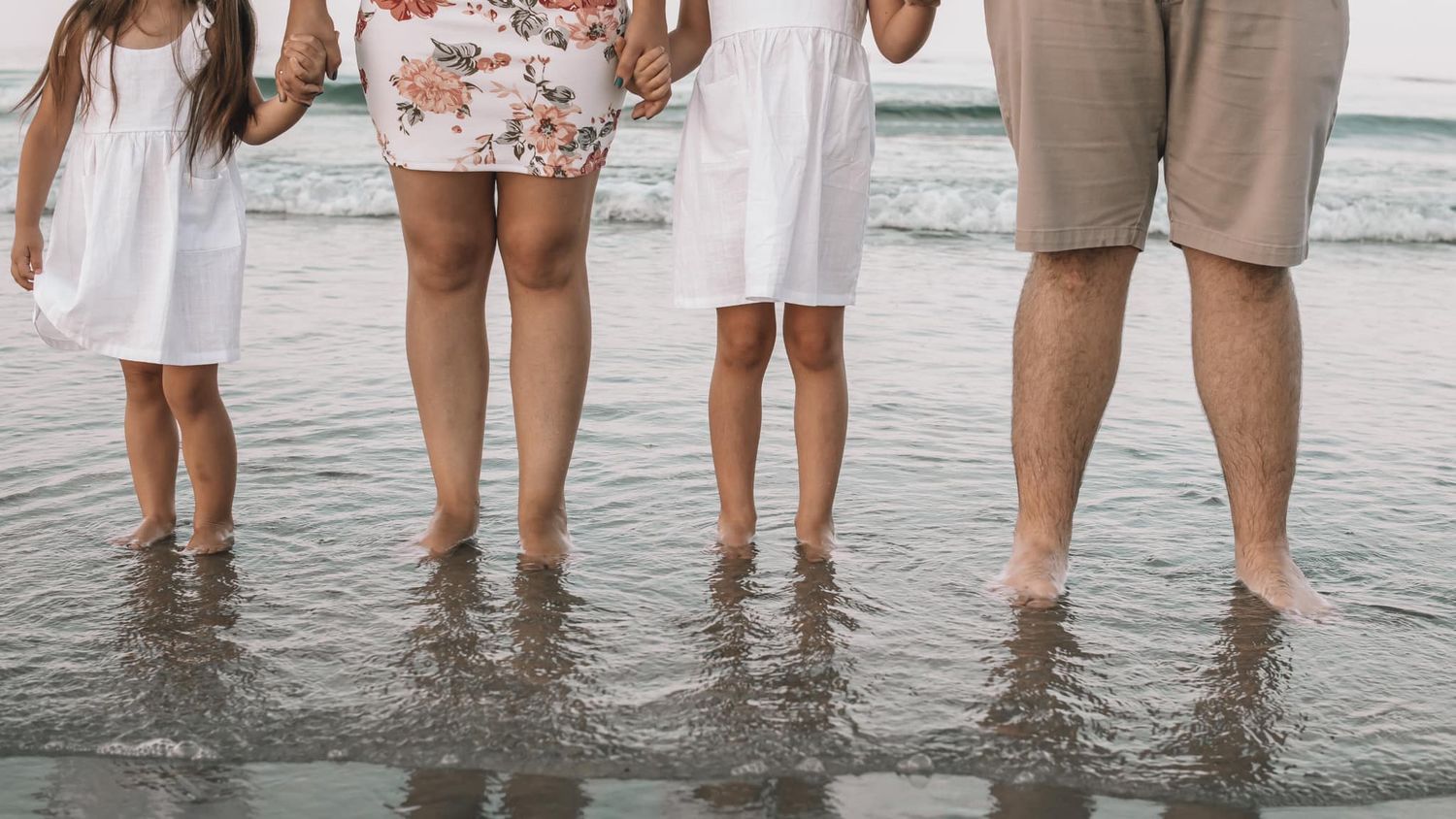 Family legs standing in shallow ocean water at the beach.