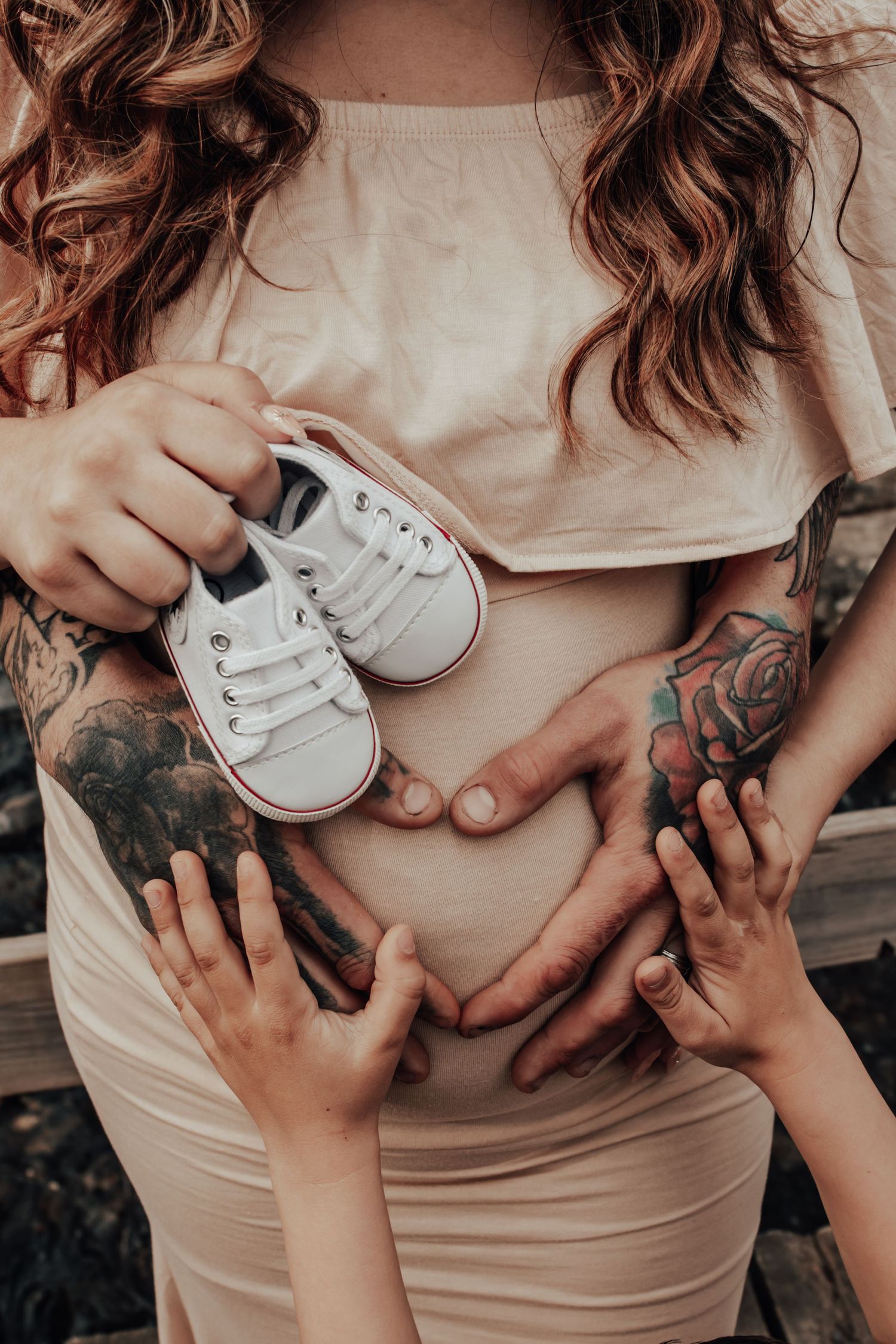 Tattooed hands hold tiny baby shoes against pregnant belly in intimate maternity photo.