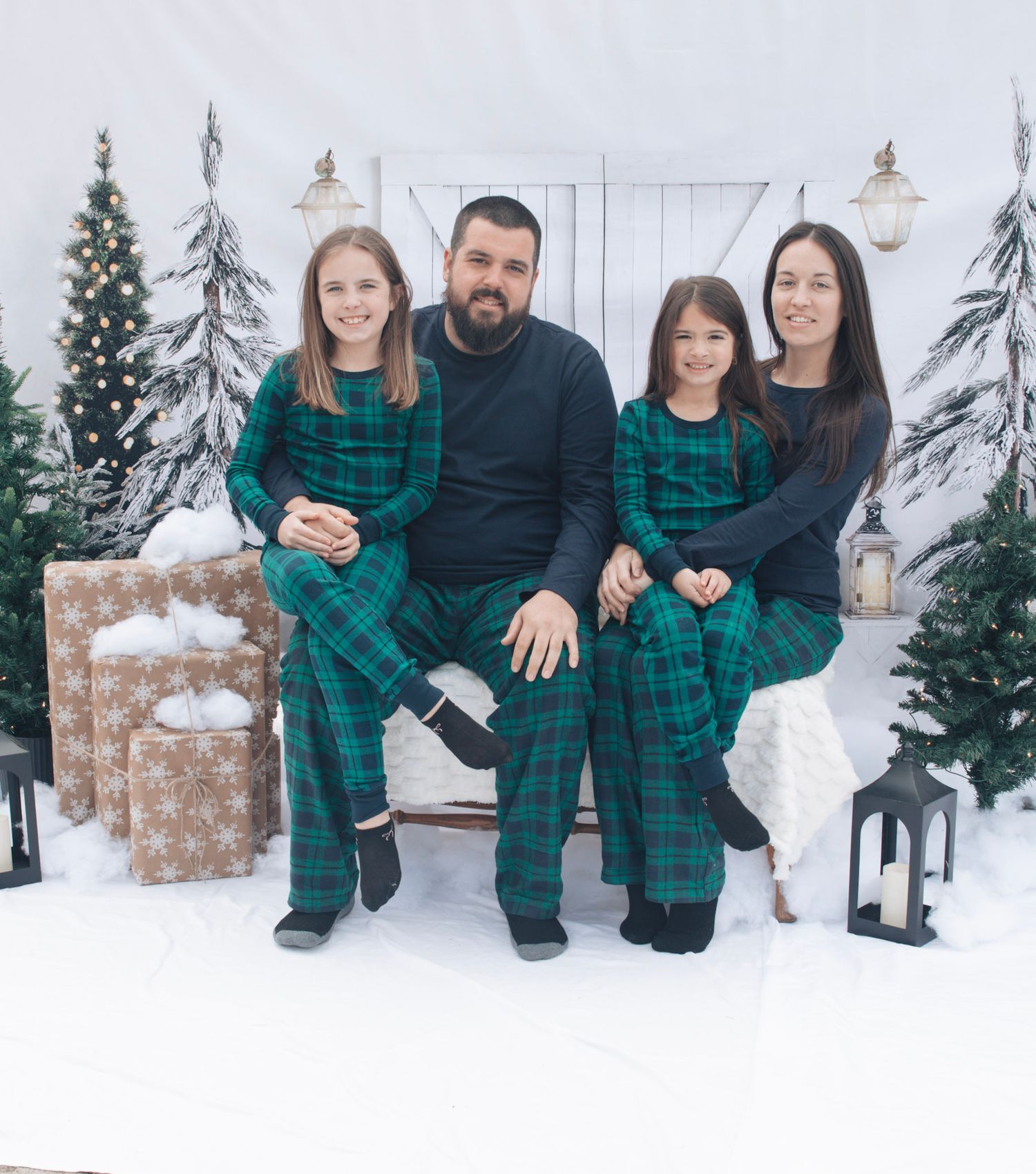 Family wearing matching green plaid outfits poses together in winter scene with Christmas trees and snow.