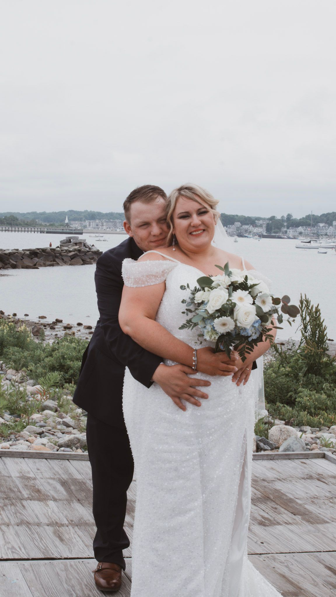 A romantic wedding photo of a couple embracing on a wooden deck overlooking a harbor with boats in the background.