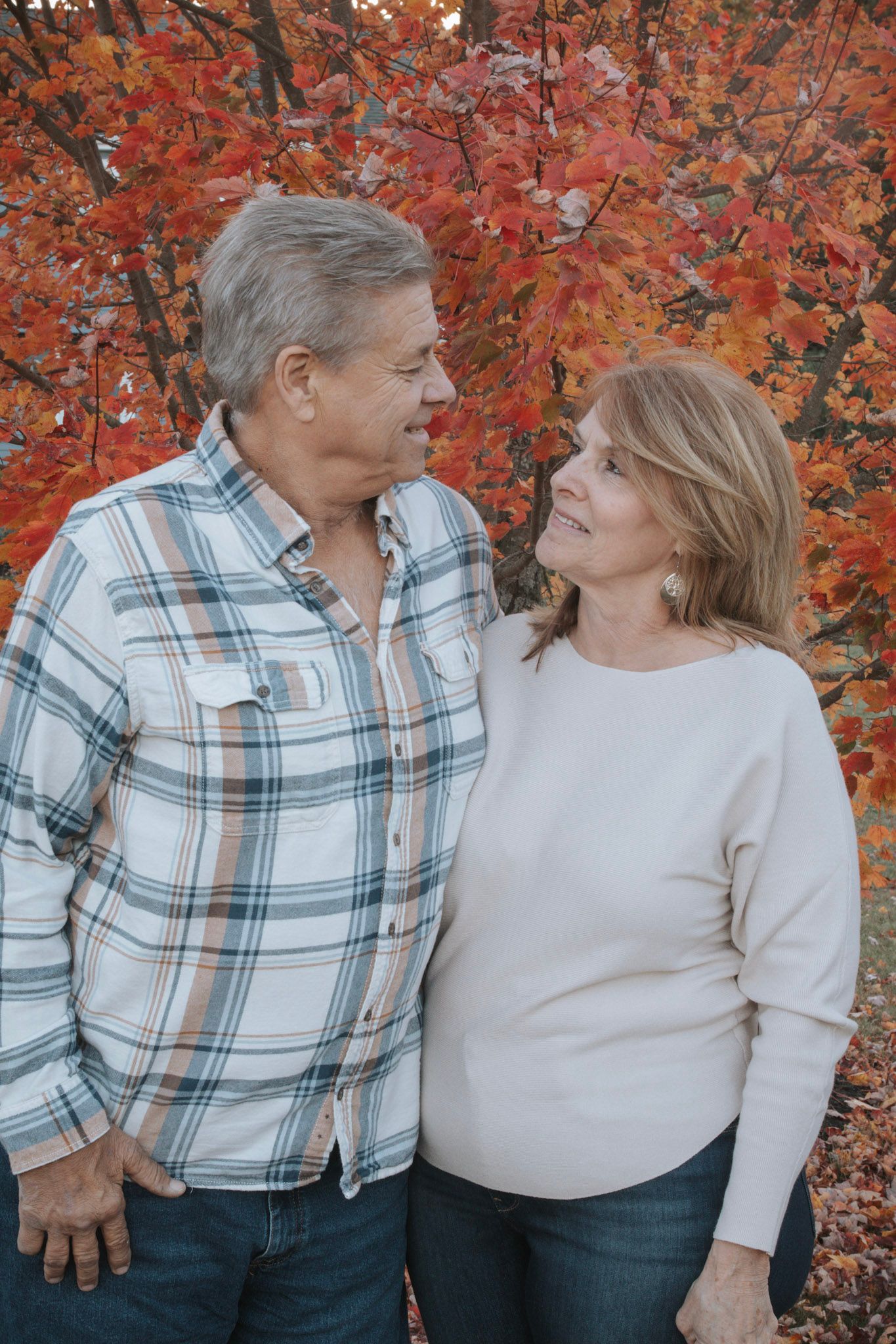 A couple wearing white and plaid embracing against a backdrop of vibrant red autumn foliage.