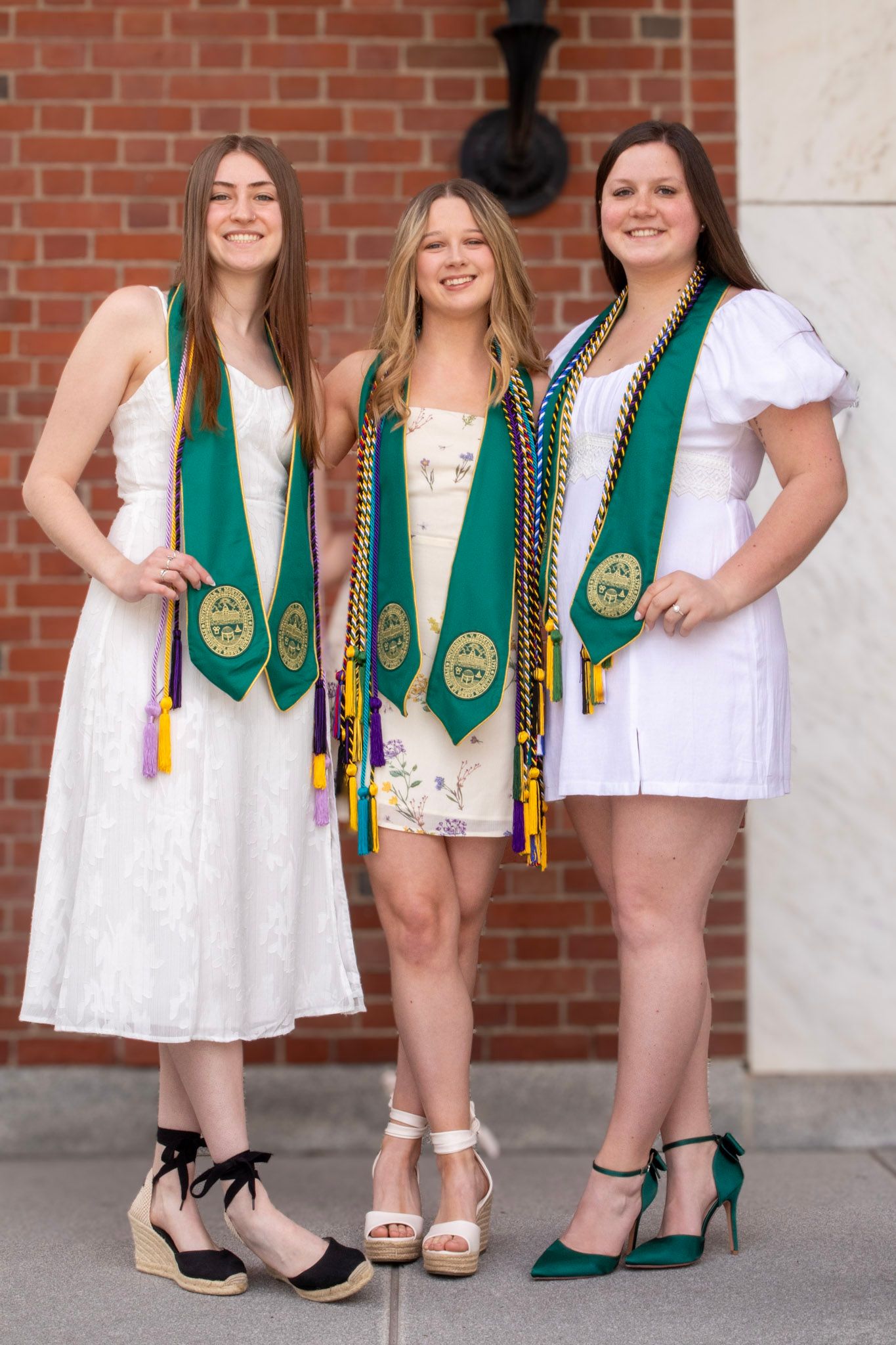 Close-up view of graduation stoles and white dresses with coordinated footwear against a brick wall background.