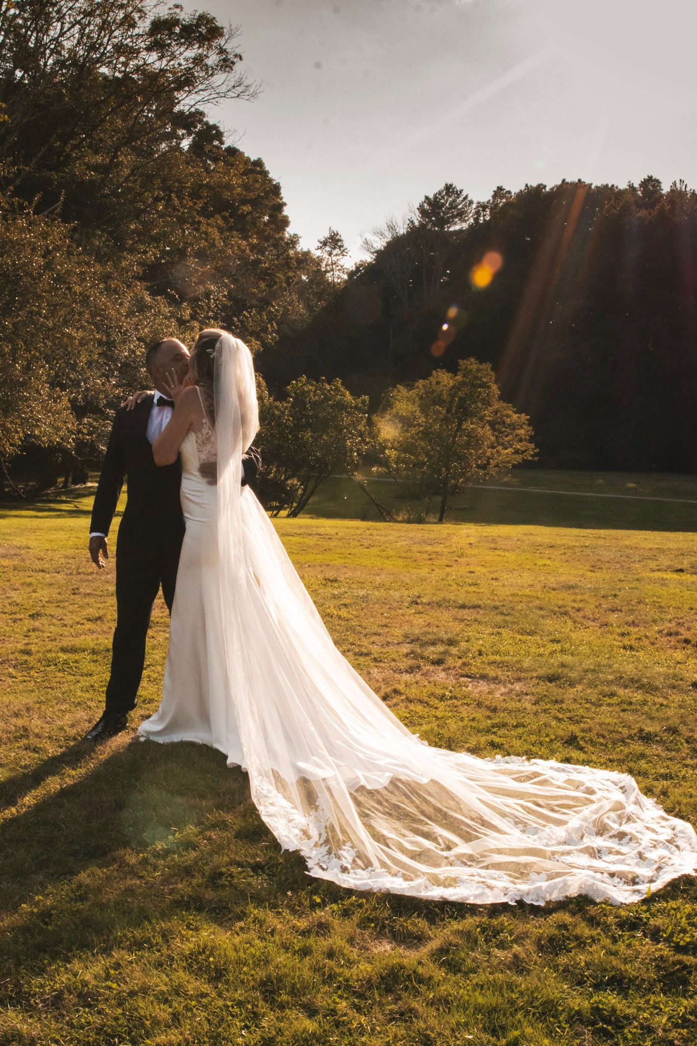 Sunset wedding portrait with bride's cathedral length veil trailing across a golden meadow.