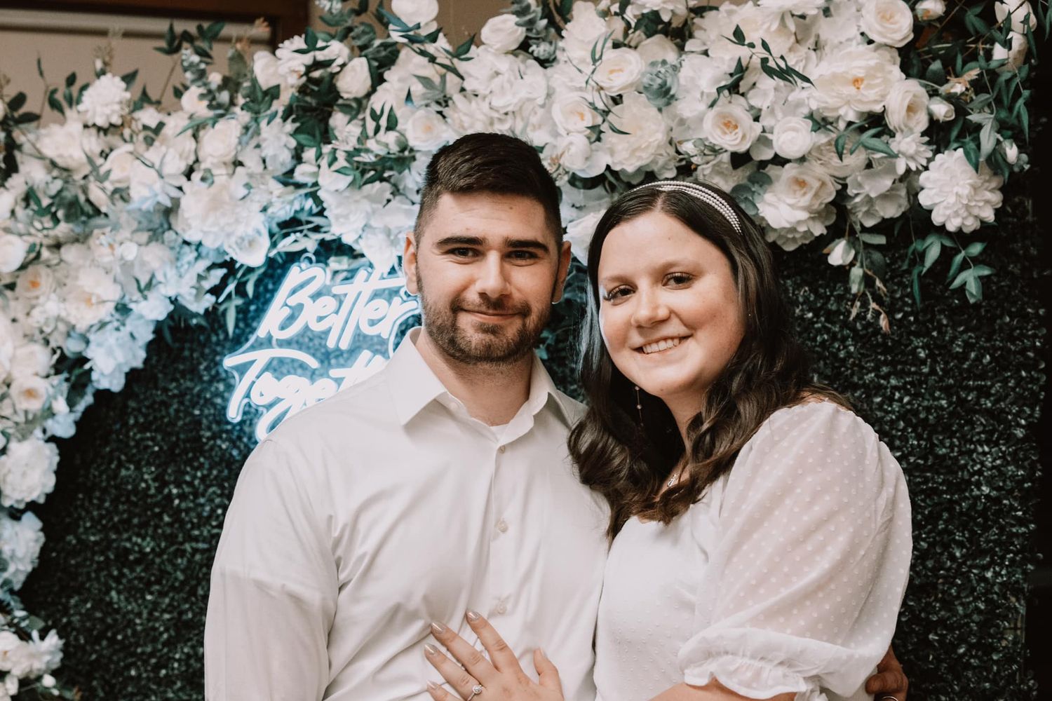 Engaged couple posing together in front of a floral wall backdrop with white flowers and neon signage.