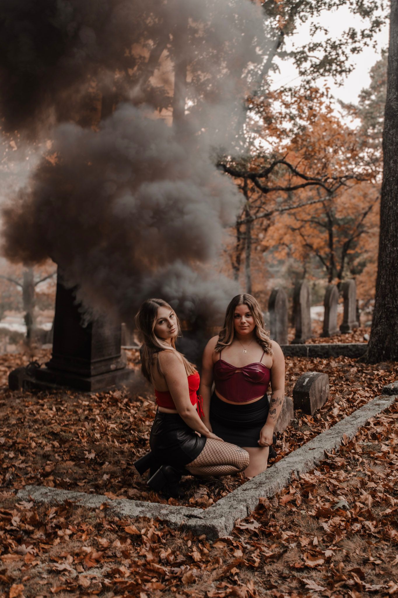Two people sit among autumn leaves with smoke effects creating a moody atmosphere in a park setting.