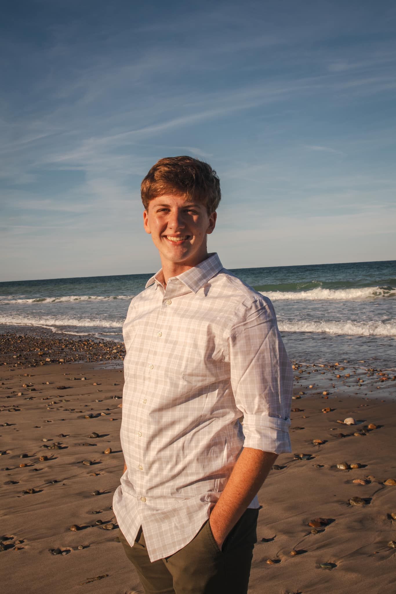 A person stands smiling on a beach wearing a white button-down shirt and khaki pants.