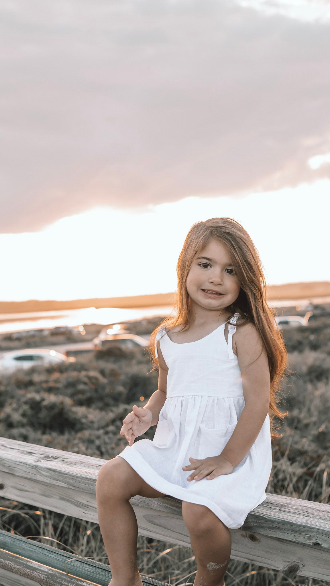 Young girl in white summer dress sitting on wooden railing during sunset at coastal overlook.