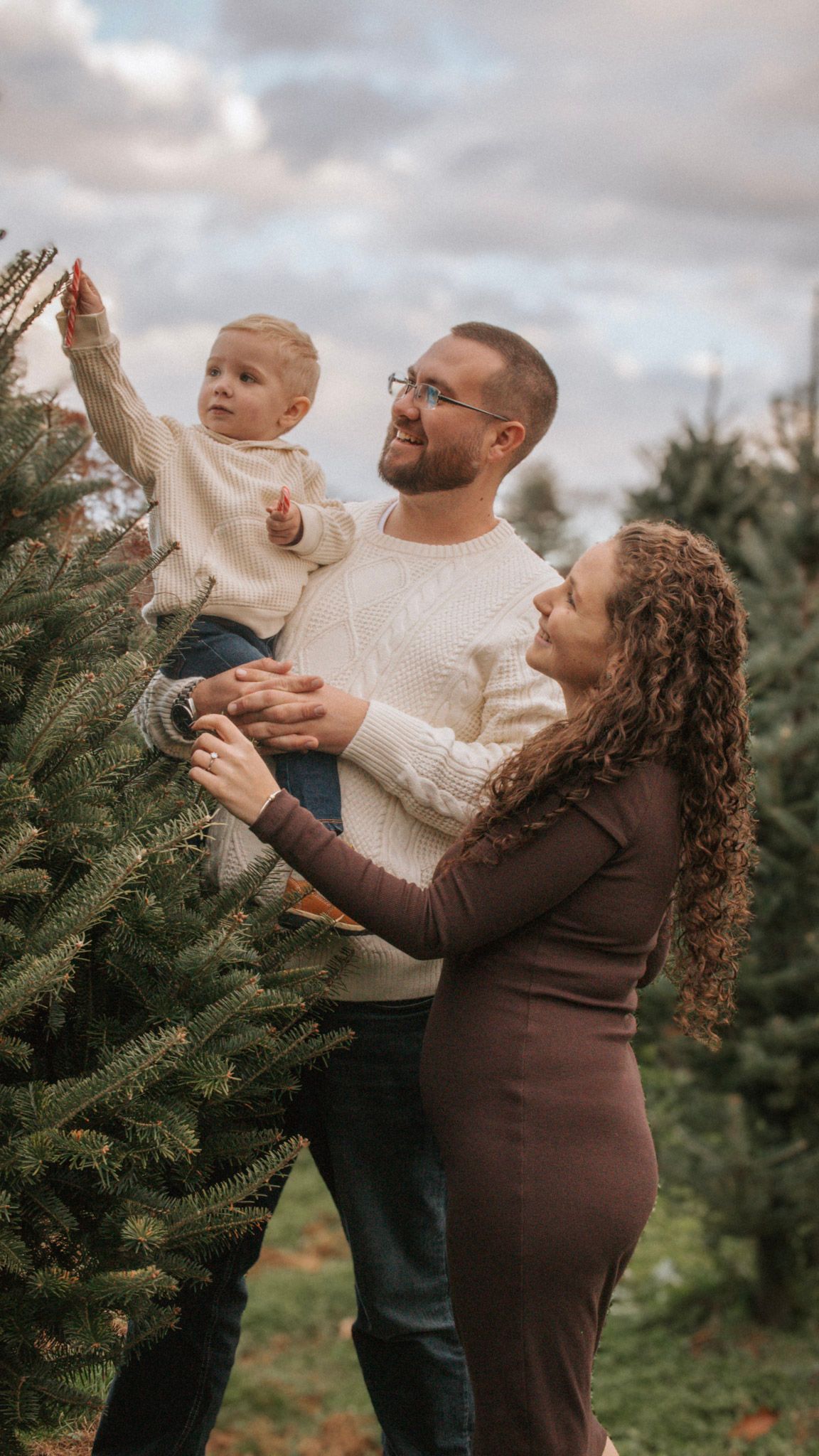 A family decorates a Christmas tree together at a tree farm on a cloudy day, all wearing cozy sweaters.
