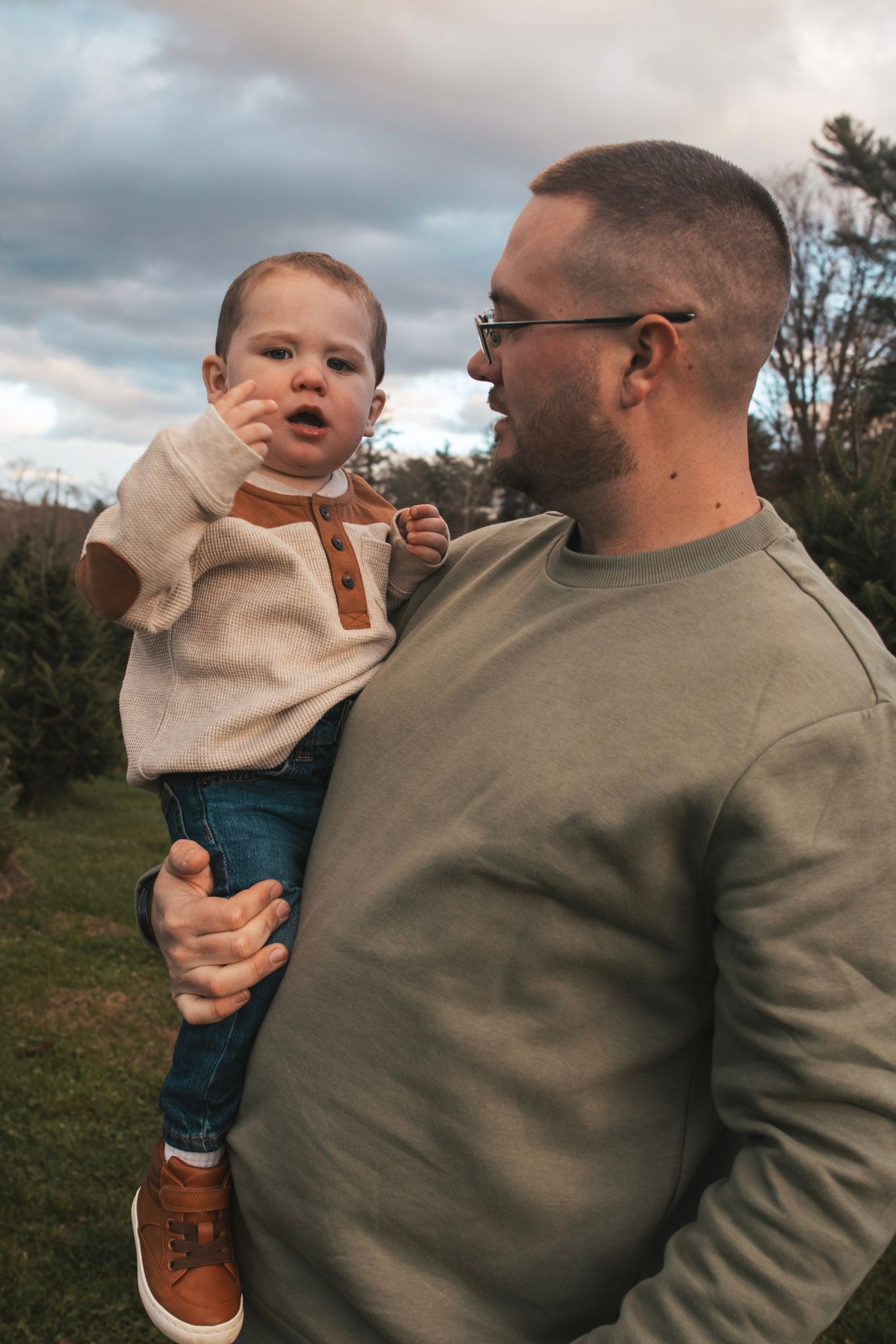 A parent holds a baby wearing a beige sweater and jeans while sharing a tender outdoor moment together.