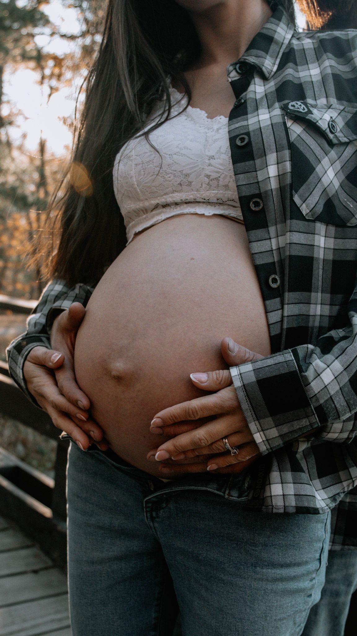 A pregnant belly is cradled in hands wearing a plaid flannel shirt and white lace top during an outdoor maternity photo shoot.