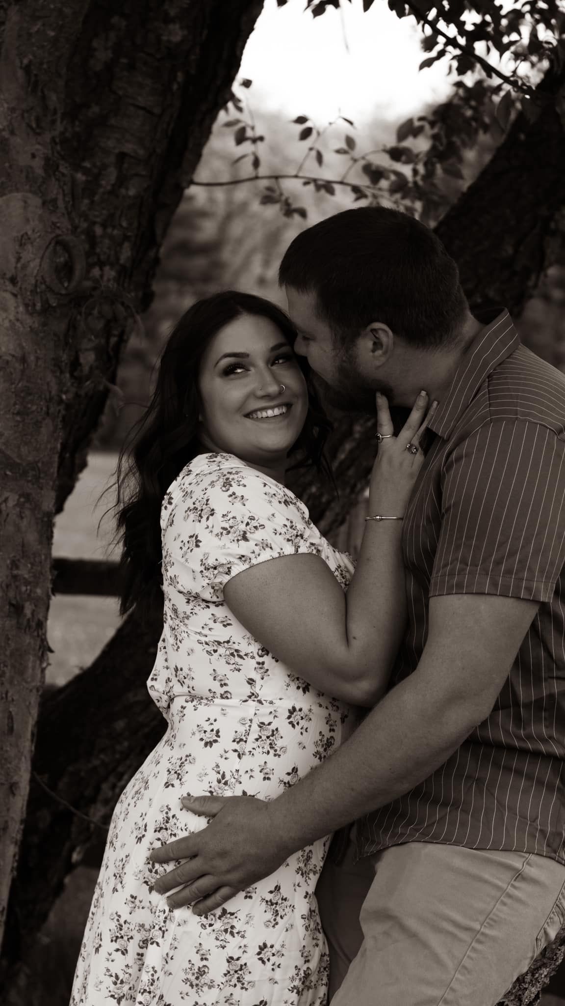 A romantic black and white portrait of a couple embracing against a tree in a floral dress and striped shirt.