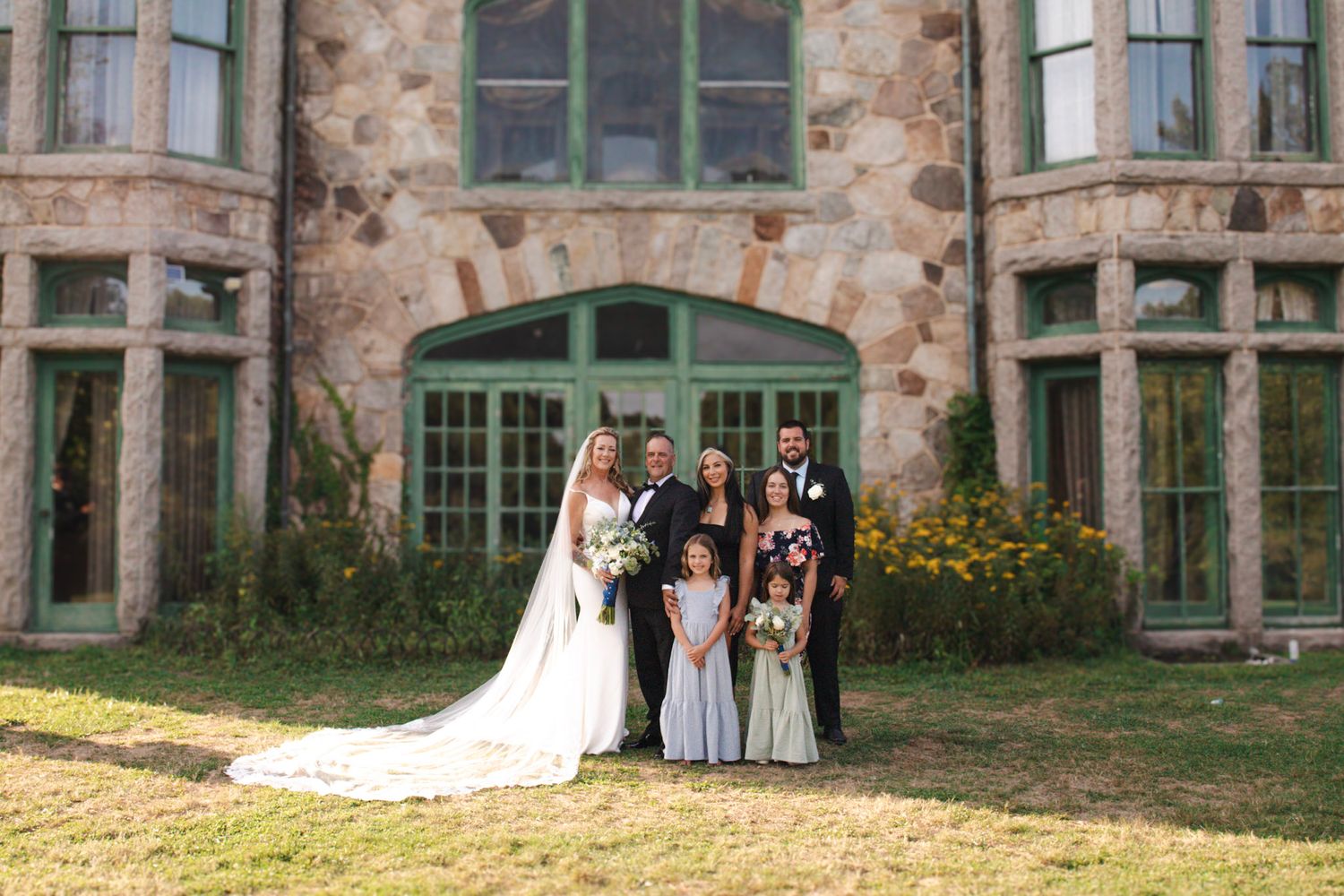 Wedding party poses in front of a stone building with bay windows and colorful garden flowers.
