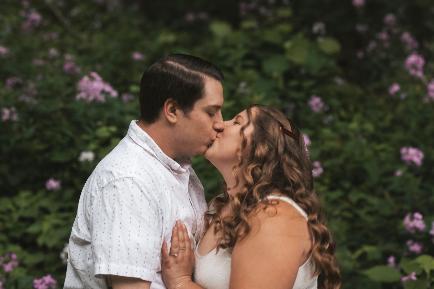 A couple shares a romantic kiss in a garden setting with pink flowers blooming in the background.