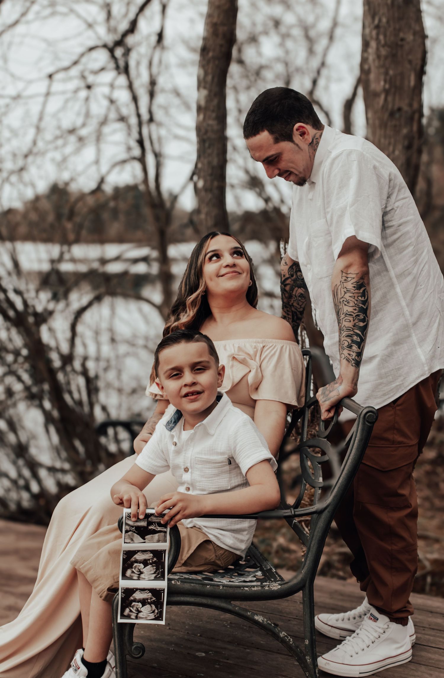 Family sits on park bench holding ultrasound photos during maternity photoshoot.