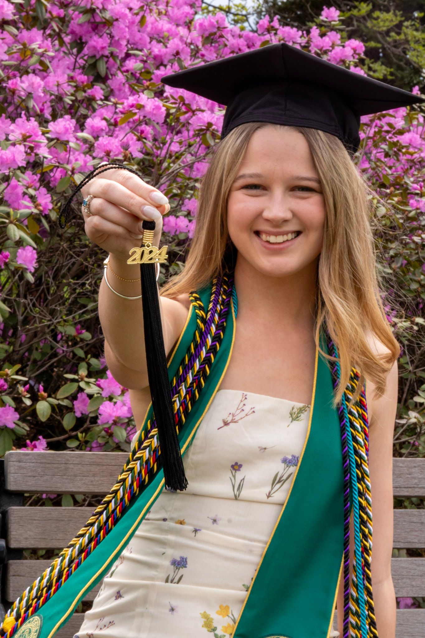 Graduate in cap and gown poses for photos in front of blooming pink azalea bushes during spring commencement ceremony.