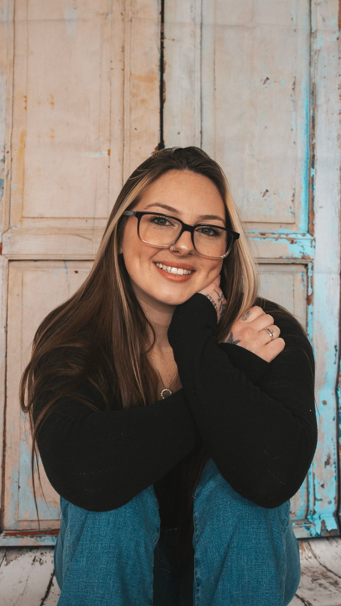 Woman in glasses and black sweater laughs joyfully against rustic wooden wall with peeling blue paint.