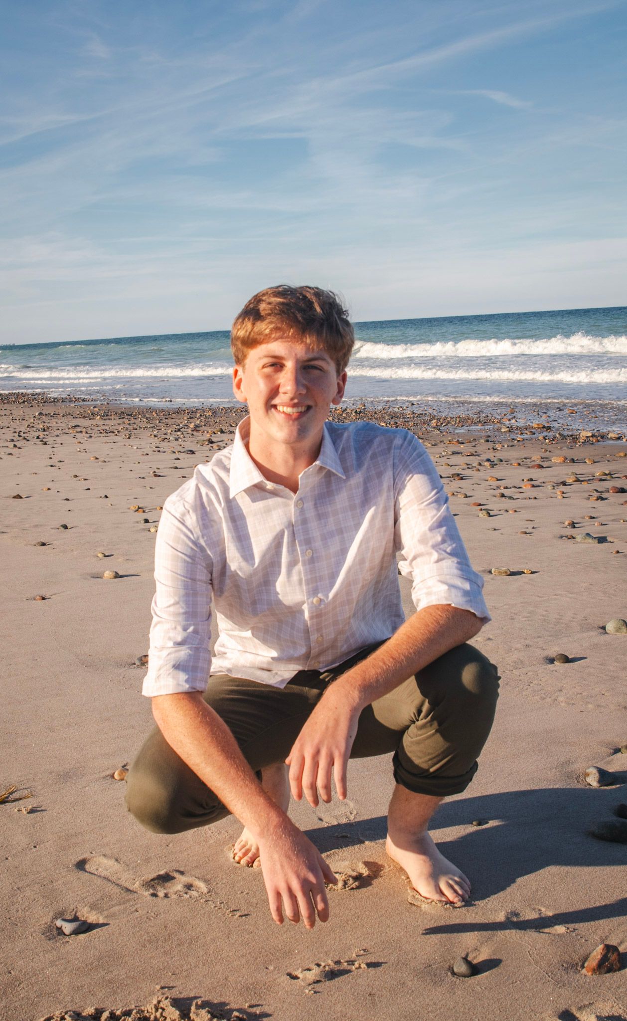 A person in a white shirt sits on a sandy beach with ocean waves and blue sky in the background.