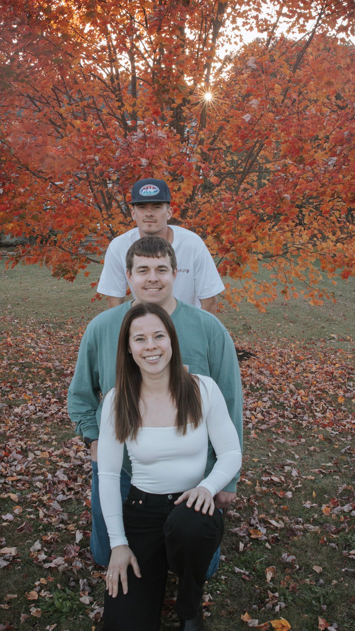 Three people stand in front of vibrant autumn maple trees with orange leaves and fallen foliage on the ground.