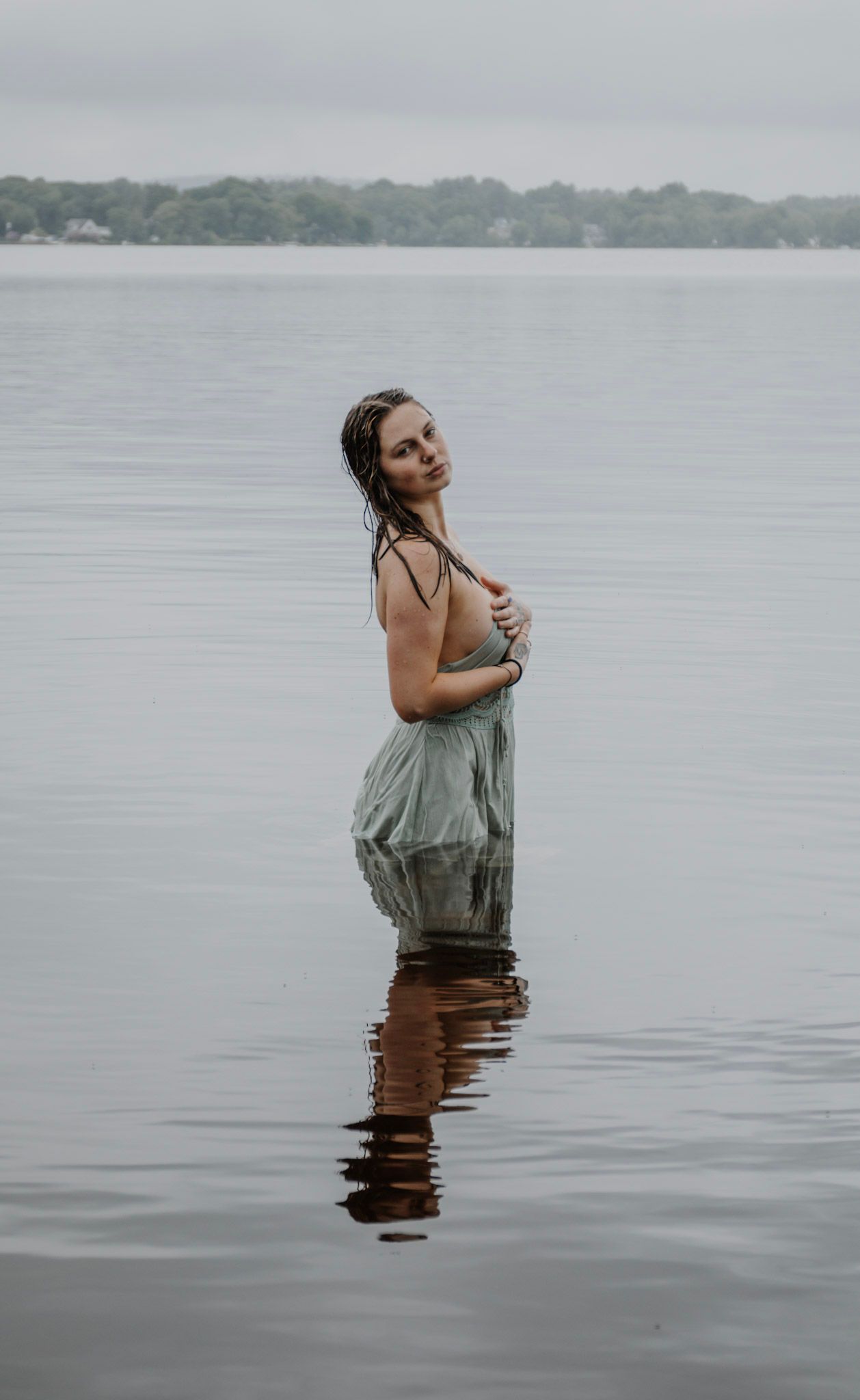 A person in a green top and brown skirt stands in calm lake waters against an overcast sky.