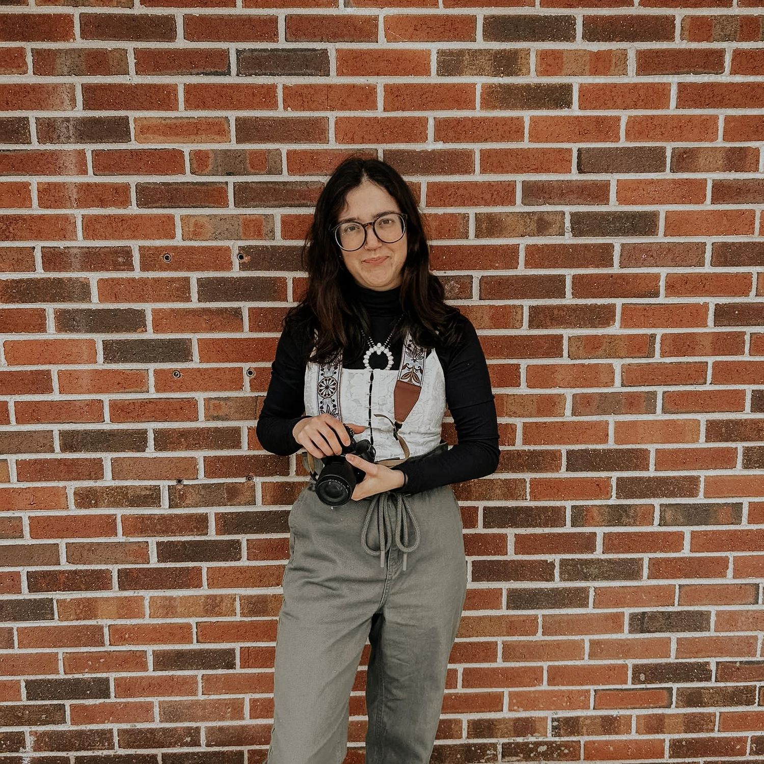 Photographer in black turtleneck and gray pants poses with camera against brick wall.