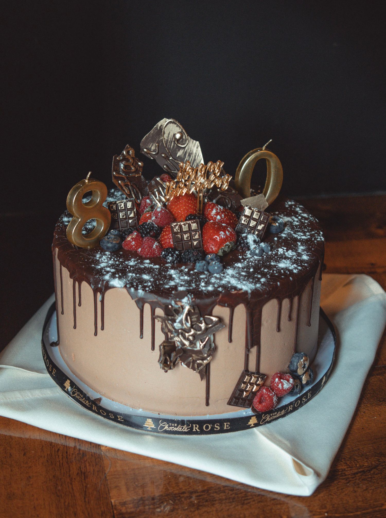An elegant chocolate drip cake decorated with berries and silver accents on a white napkin.