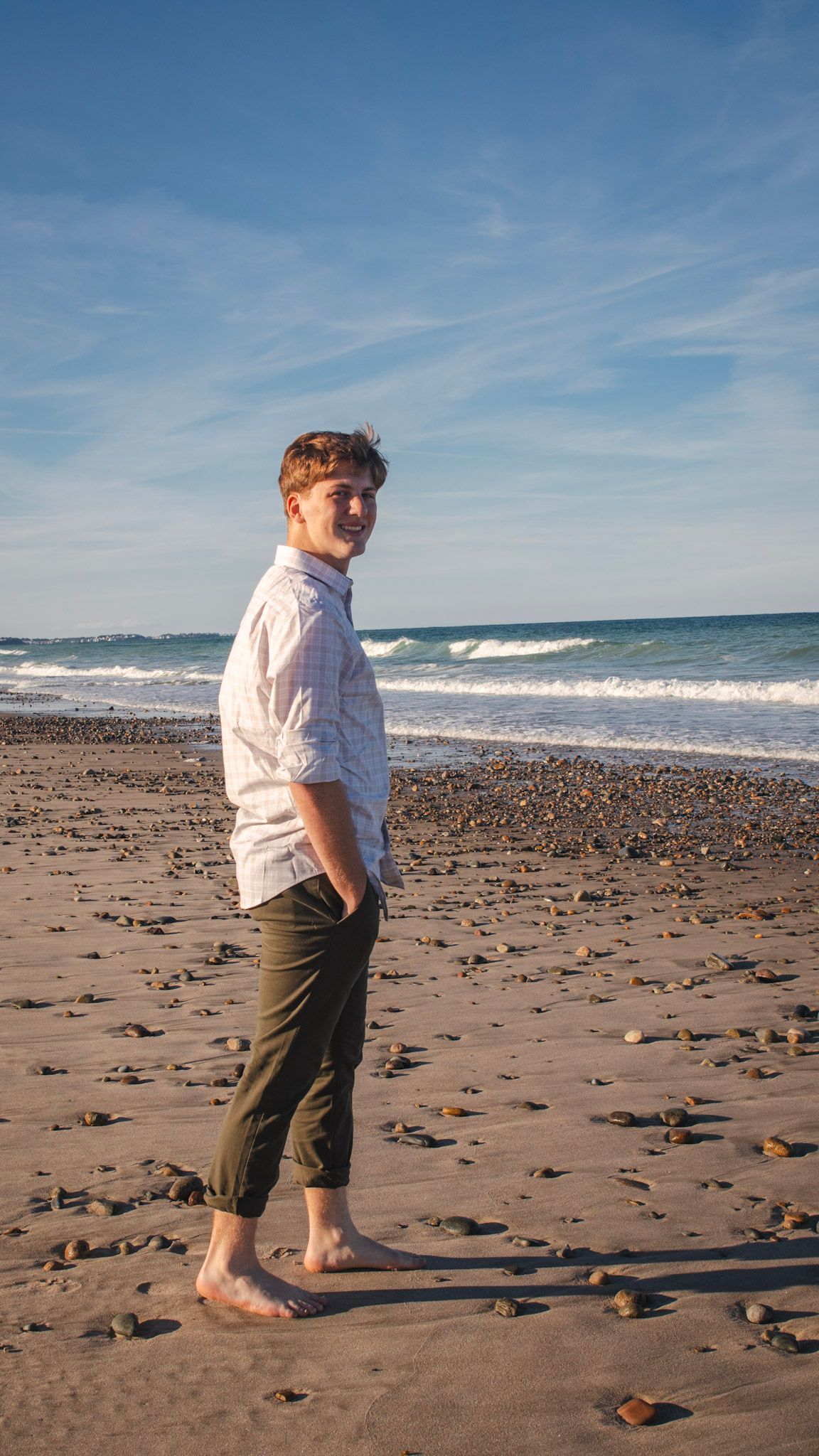A barefoot person in a cap and shorts walks along a rocky beach with ocean waves and blue sky in the background.