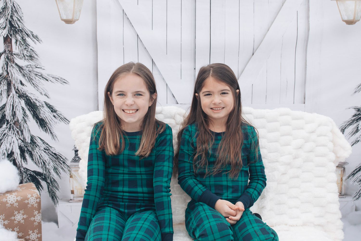 Two people in matching green plaid shirts pose together in a snowy winter setting with evergreen trees.