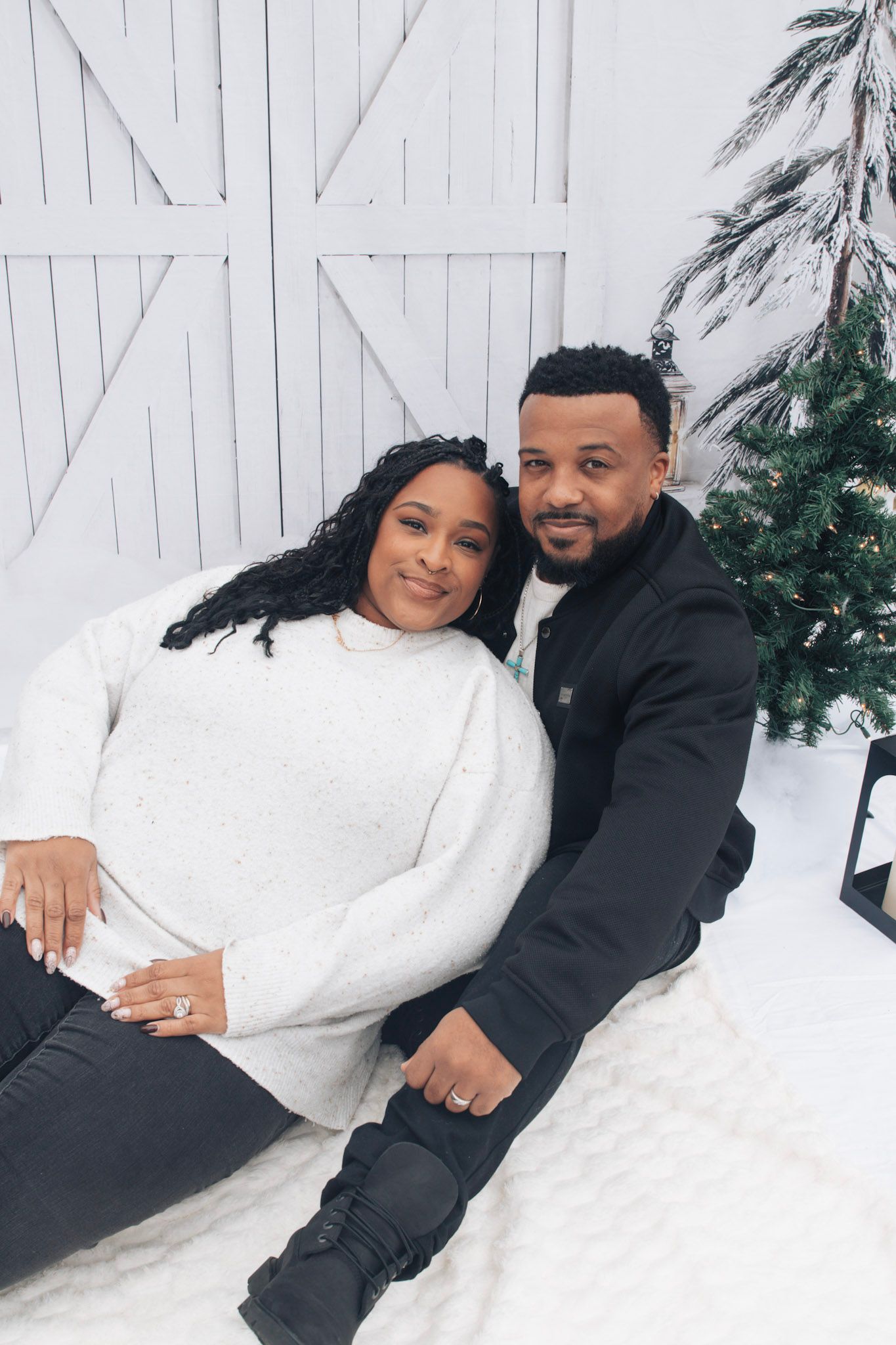 A couple wearing black and white outfits poses together in a winter scene with snow and evergreen trees.