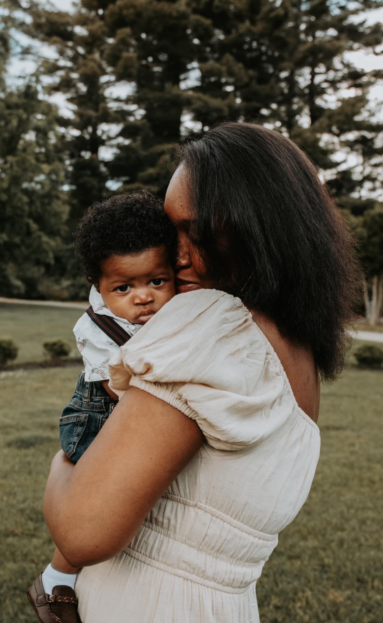 A parent and child share embraces outdoors in a grassy field with pine trees.