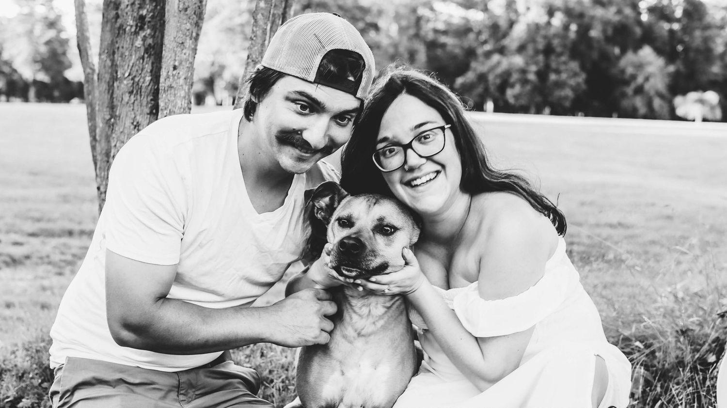Happy couple poses with their small dog in a sunny park setting.