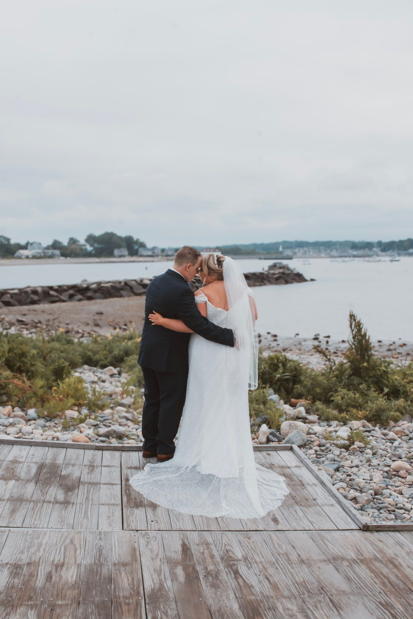 Romantic wedding couple embraces on a wooden boardwalk overlooking a rocky beach coastline.