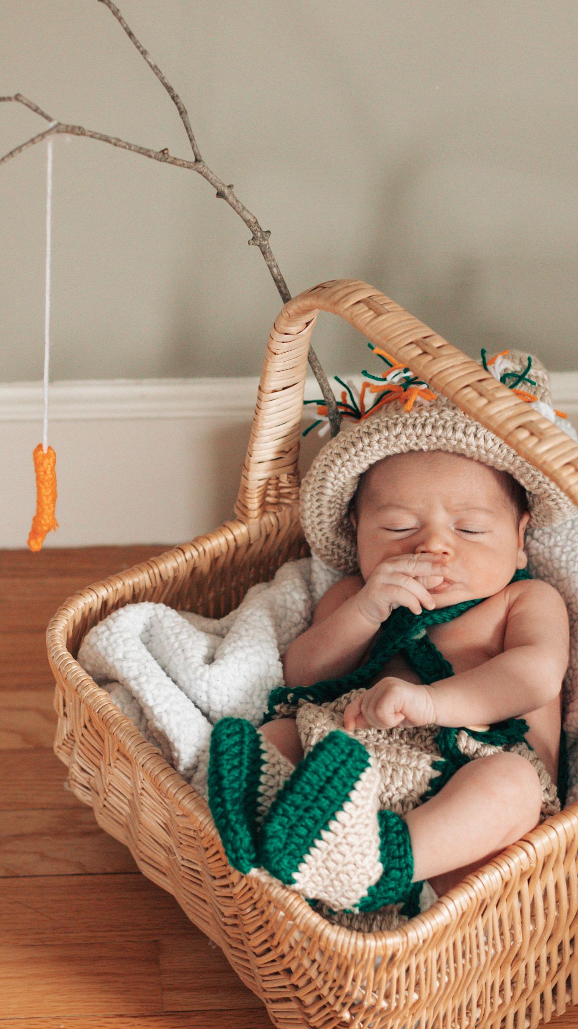 Sleeping newborn in wicker basket wearing white and green knitted outfit with carrot-themed accessories.