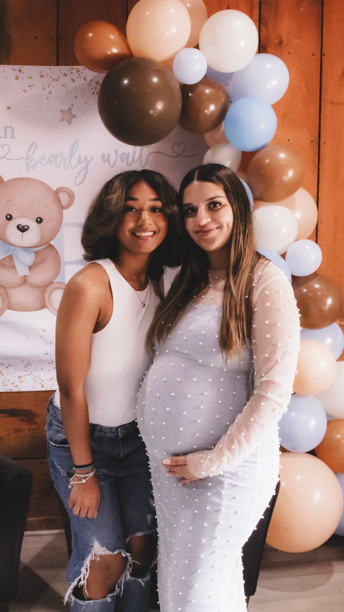 Two friends pose together at a baby shower with brown and white balloon decorations and teddy bear backdrop.