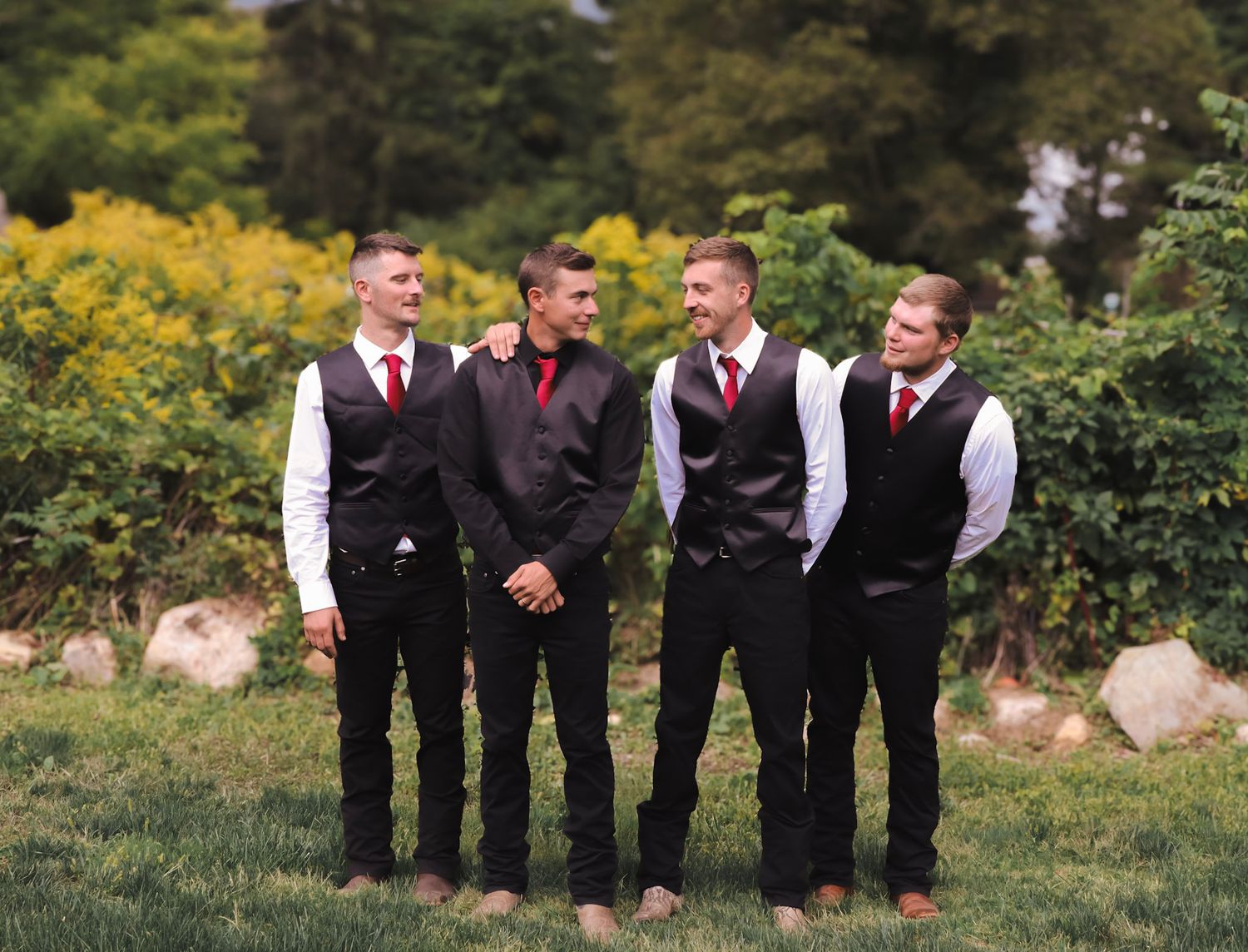 Groomsmen in black suits and red ties stand in formation outdoors against a natural backdrop.