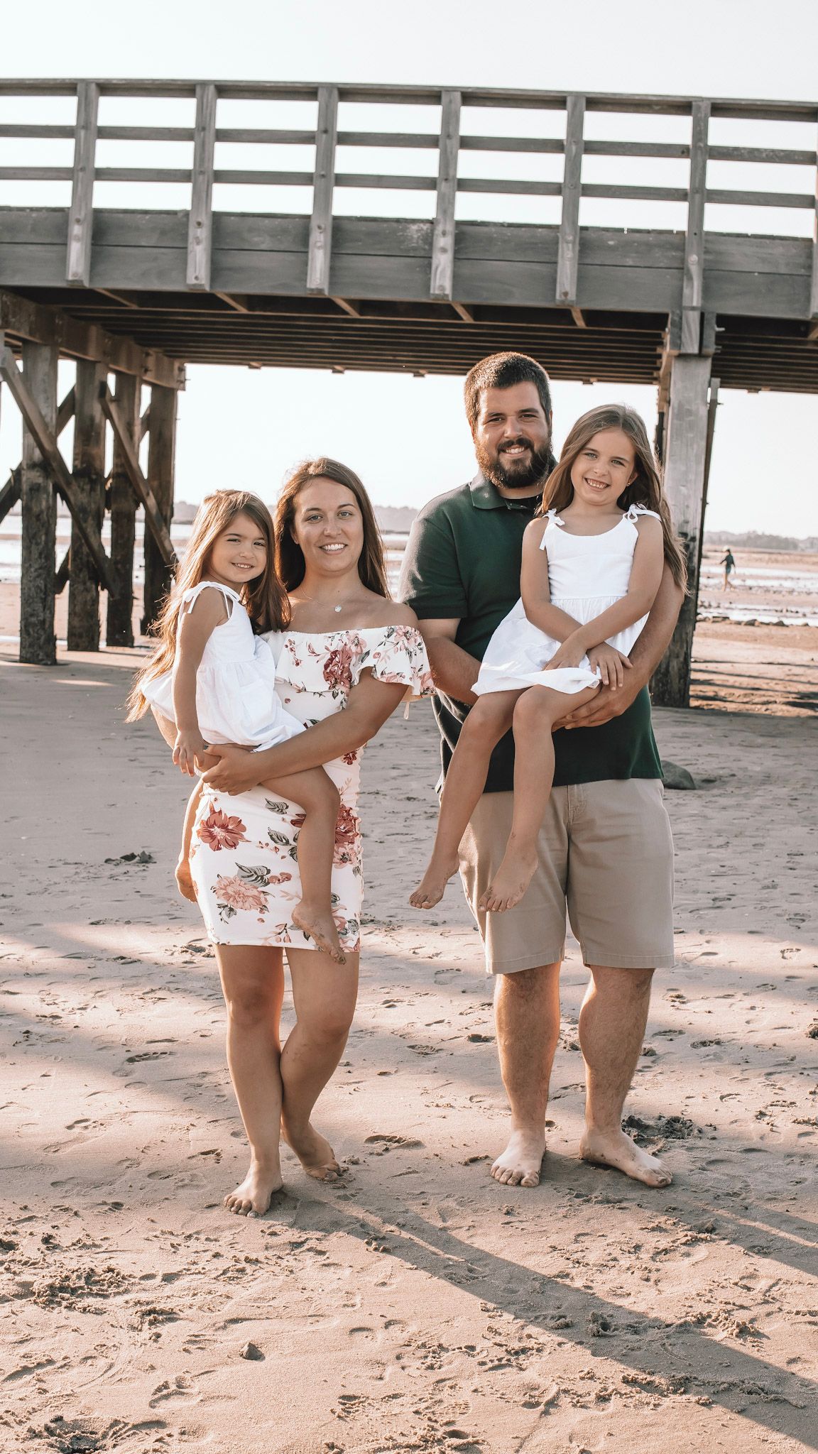 Family poses playfully under a wooden pier on a sandy beach during a casual photoshoot.