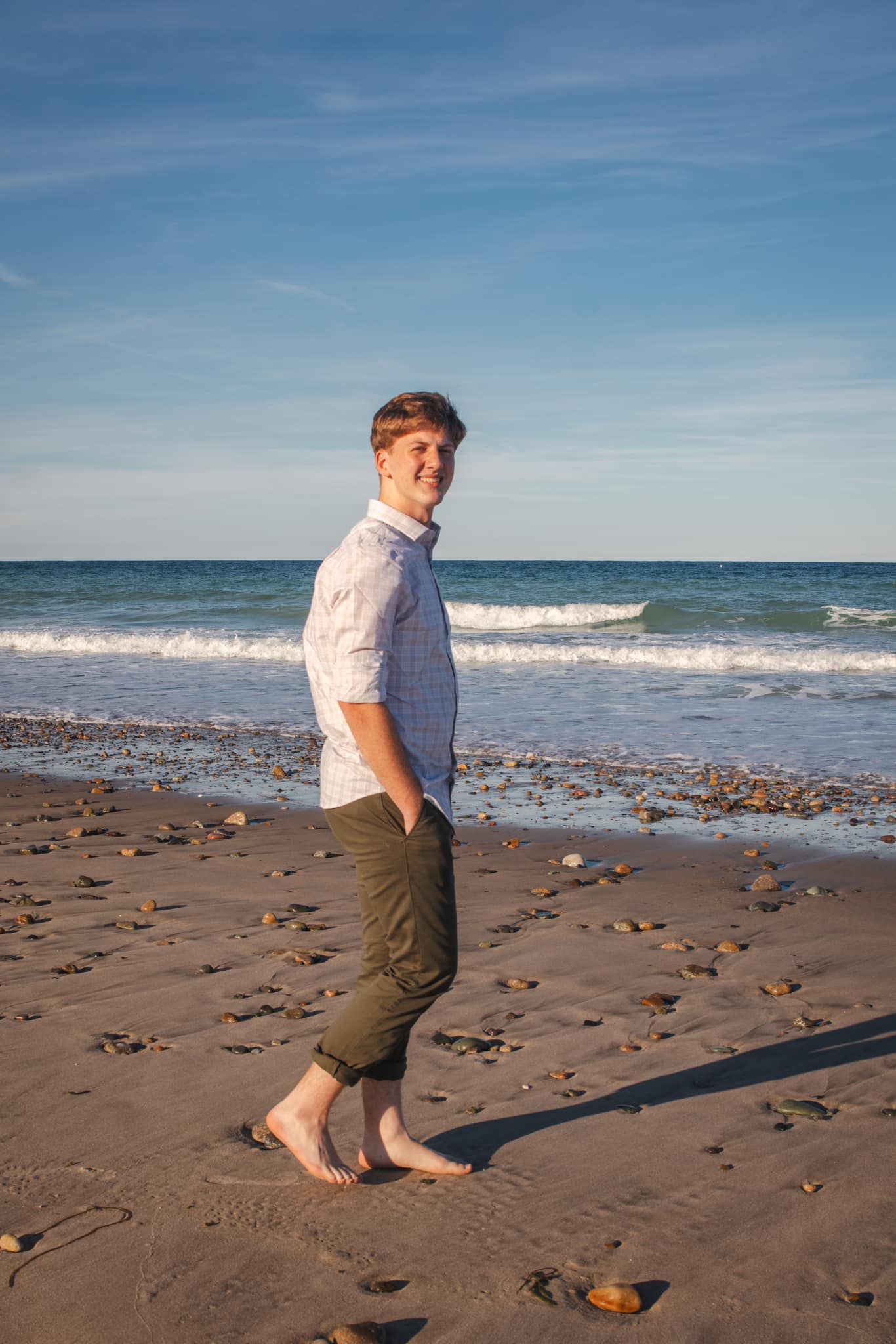 A person in a white shirt and rolled-up pants walks barefoot along a rocky beach shoreline.
