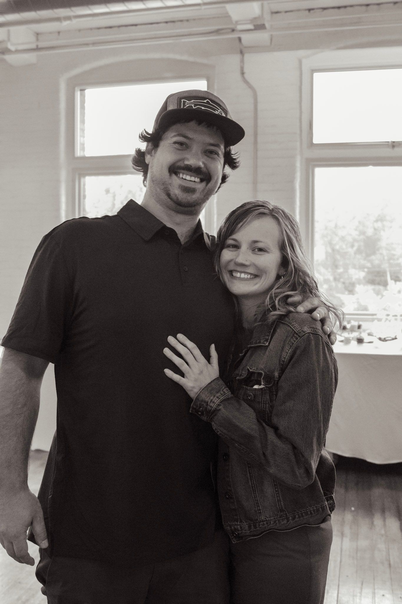 A black and white portrait of a couple embracing in a bright indoor space with windows.