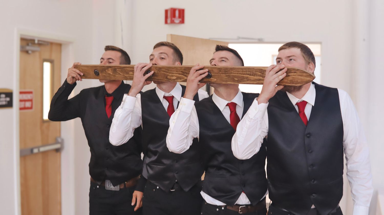 Groomsmen in matching black vests and red ties pose playfully while drinking from champagne bottles in a hallway.