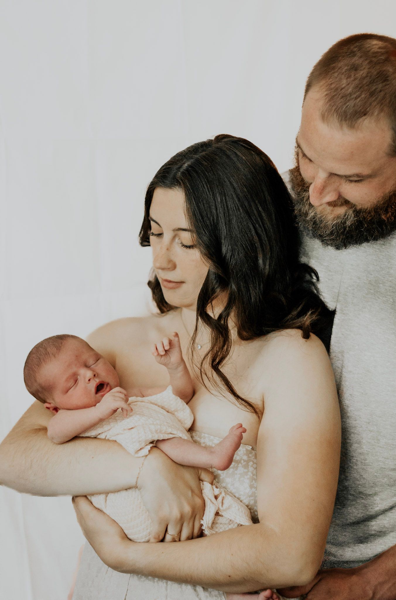 A tender family moment as parents hold their newborn baby against a white backdrop in an intimate portrait.