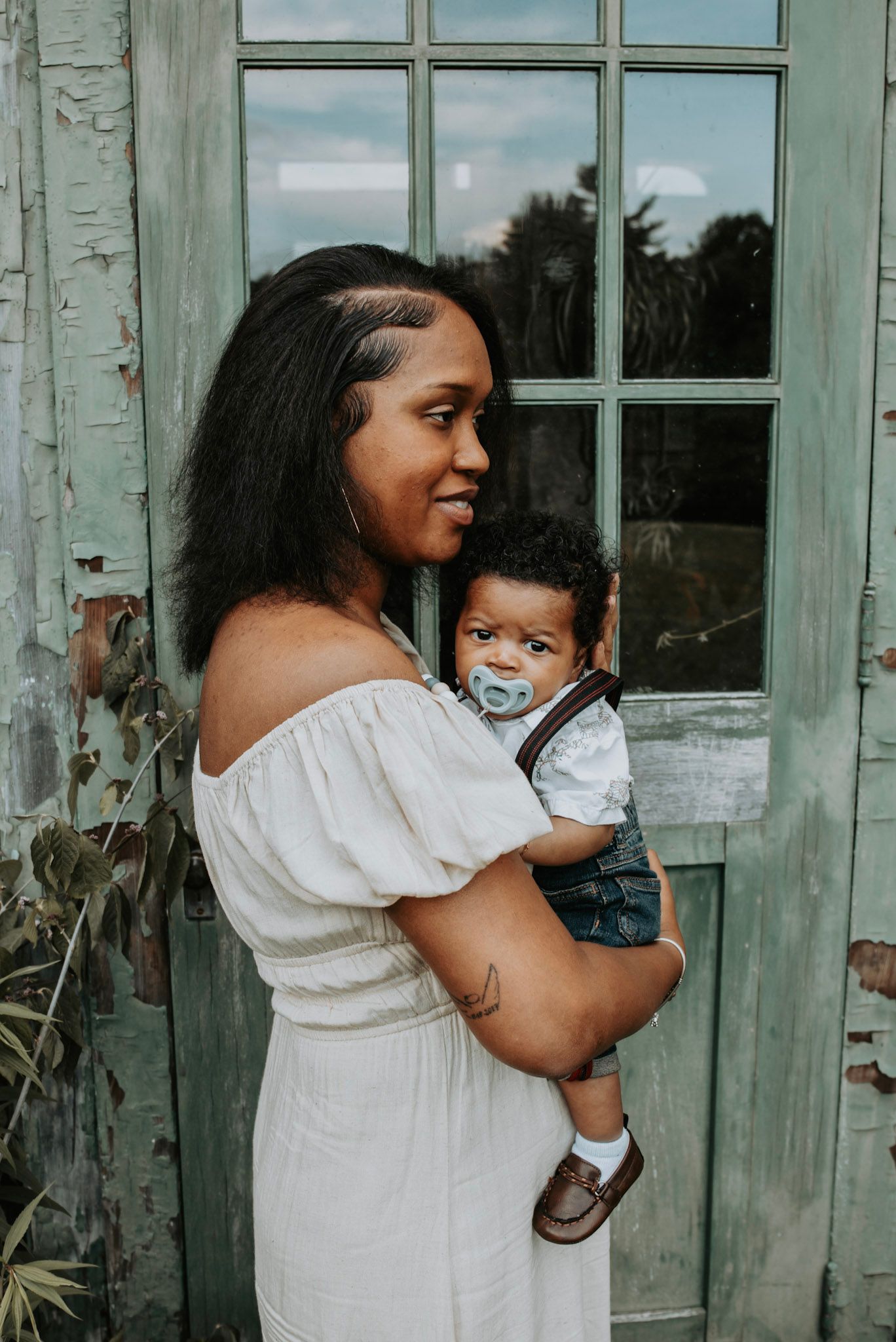 A tender moment captured of a mother in a white dress holding her baby in front of a weathered turquoise door.