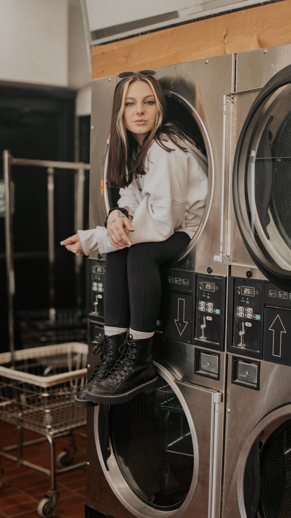 Person wearing black pants and boots sits on industrial washing machines in a laundromat setting.