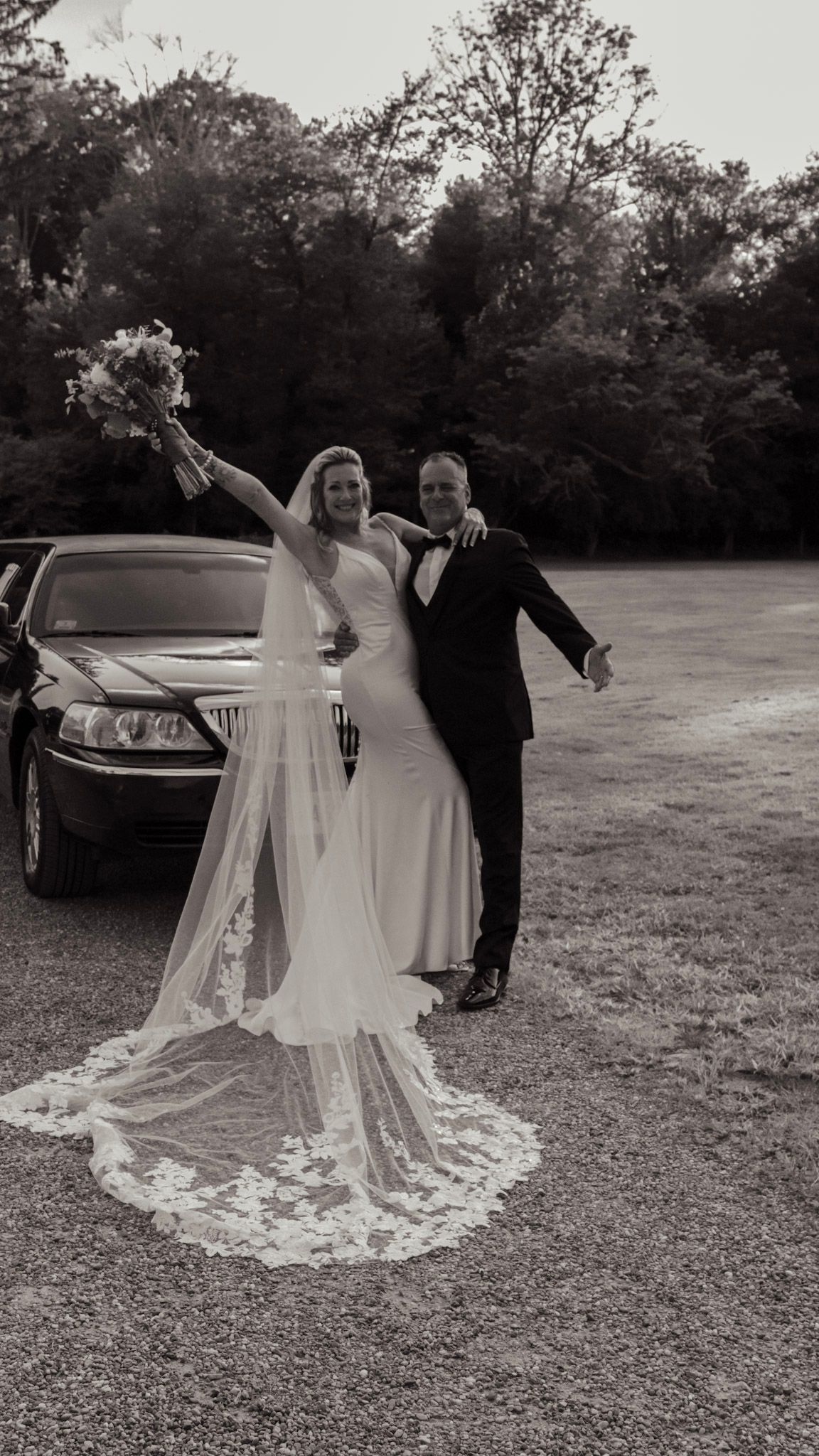 A wedding couple poses outdoors in black and white with a flowing lace veil creating dramatic movement.