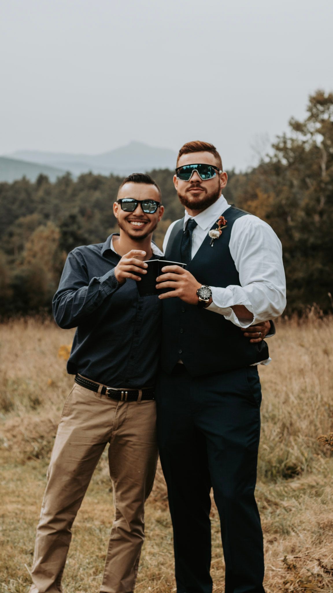Two friends in sunglasses and formal wear sharing drinks and laughs in an outdoor field setting.