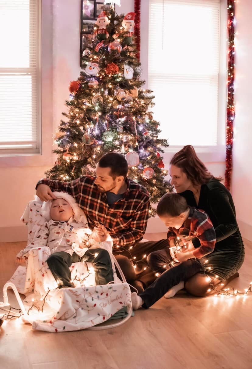 Family sits together on floor with Christmas lights and decorated tree in cozy holiday scene.