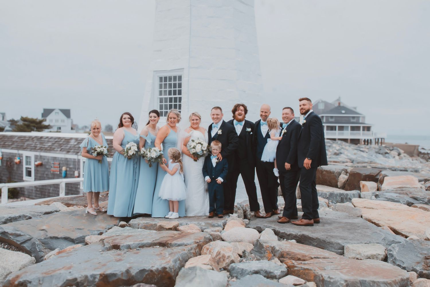 Wedding party poses together on a rocky beach jetty at sunset with ocean waves in the background.
