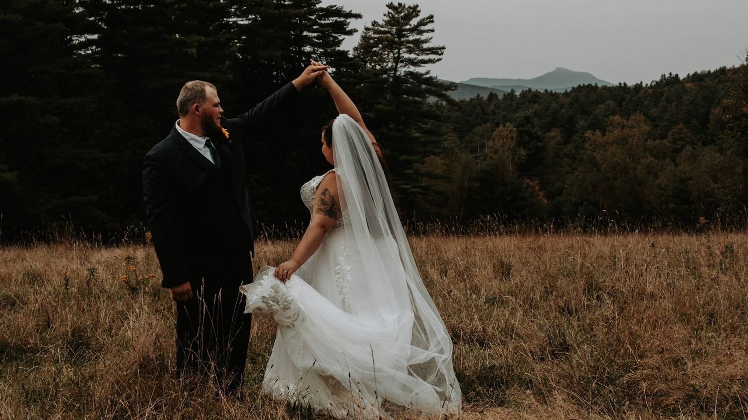 A couple in wedding attire walks through a golden field at sunset with their veil flowing in the breeze.