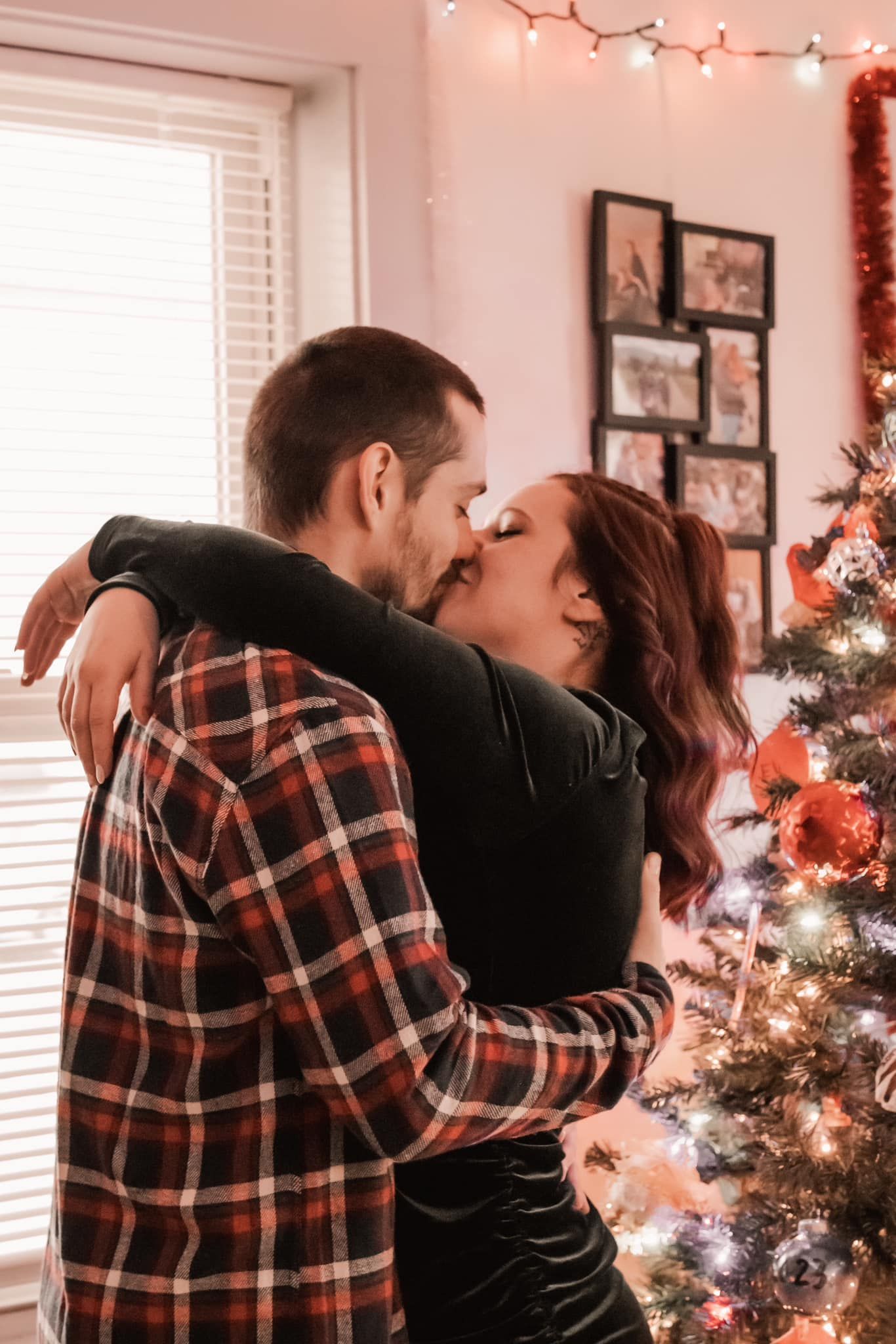 Couple sharing a romantic kiss by a decorated Christmas tree with string lights in the background.