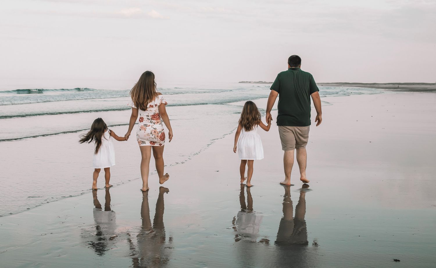 Family holding hands while walking along a misty beach at sunset, their reflections visible in the wet sand.